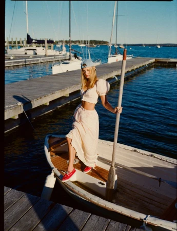 A blonde woman in a matching set stands on a small boat at a dock on a sunny day, posing with an oar and wearing a baseball cap.