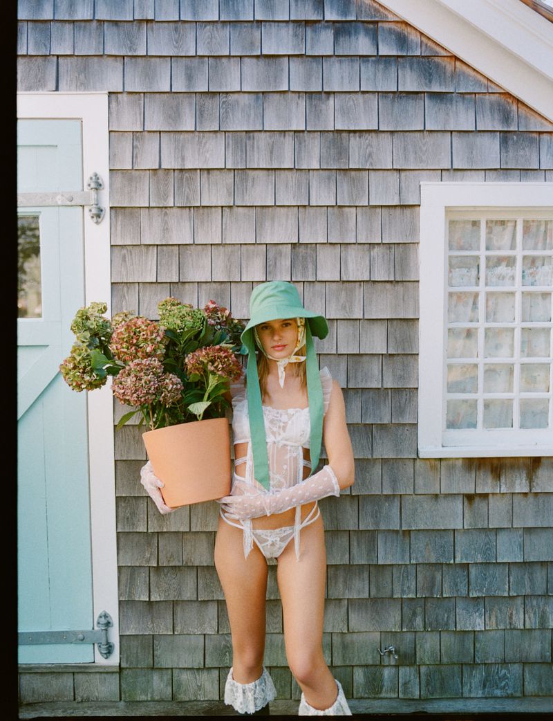 A woman in lingerie, gloves, and a bonnet stands outdoors holding a potted plant in front of a cedar-shingled house with a window.