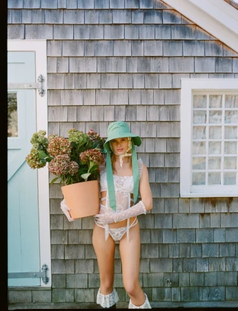 A woman in lingerie, gloves, and a bonnet stands outdoors holding a potted plant in front of a cedar-shingled house with a window.