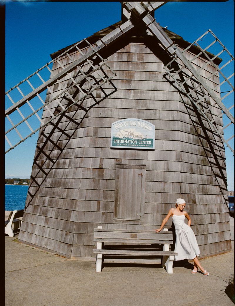 Woman in a dress poses on a bench in front of the Sag Harbor Whaling and Historical Museum Windmill under a clear blue sky.