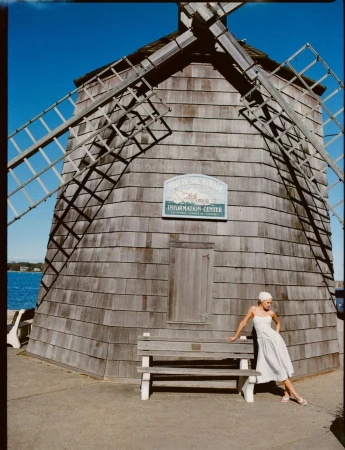 Woman in a dress poses on a bench in front of the Sag Harbor Whaling and Historical Museum Windmill under a clear blue sky.