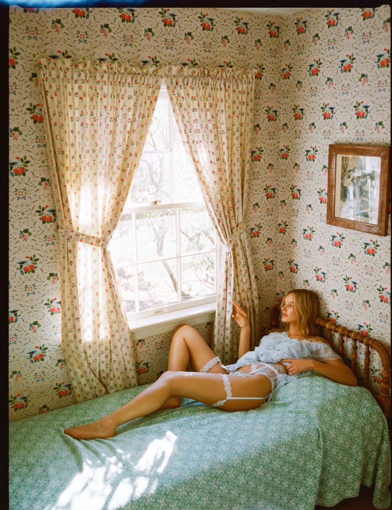 Young woman in lingerie lounges on a bed in an indoor setting with floral wallpaper and natural light from a nearby window.