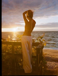 Model standing on a wooden balcony overlooking the ocean at sunset, wearing white trousers and a bikini top with hands in hair.