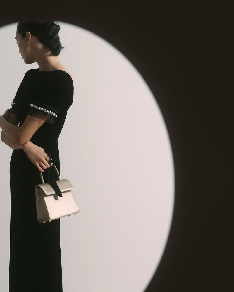 Elegant woman in black dress holding a neutral handbag, spotlight effect, fashion editorial photography, indoor environment.