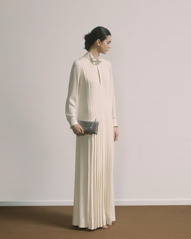 A woman in an elegant long-sleeve gown poses indoors with a gray clutch, looking left against a beige wall and brown flooring.