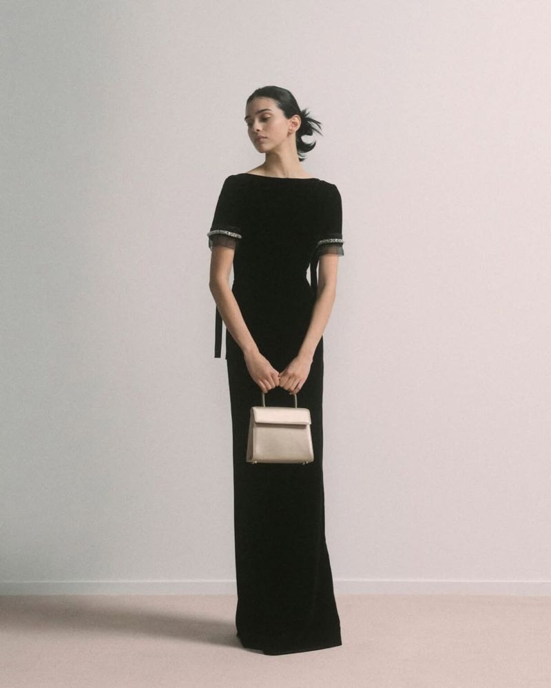 Elegant woman in black velvet gown holds gold purse against a neutral backdrop, fashion photography, studio shot.