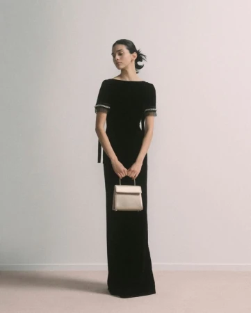 Elegant woman in black velvet gown holds gold purse against a neutral backdrop, fashion photography, studio shot.
