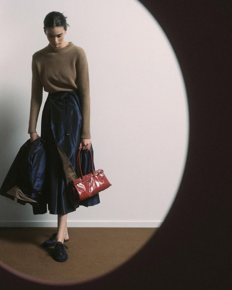 Fashion portrait of a woman in a brown sweater and blue skirt, holding a jacket and red purse, in a photo studio with a circular frame.