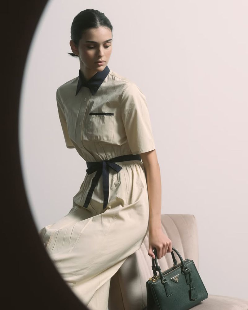 Indoor shot of a model sitting and holding a Prada handbag, wearing a beige dress with a dark blue belt, and a dark blue collar.