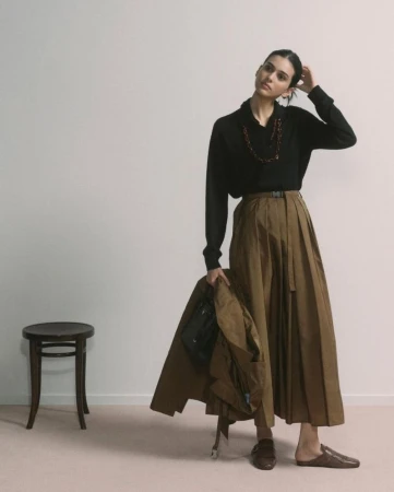 A woman in a black sweater and wide-legged brown skirt holds a jacket. The shot is set in a photo studio against a white background.