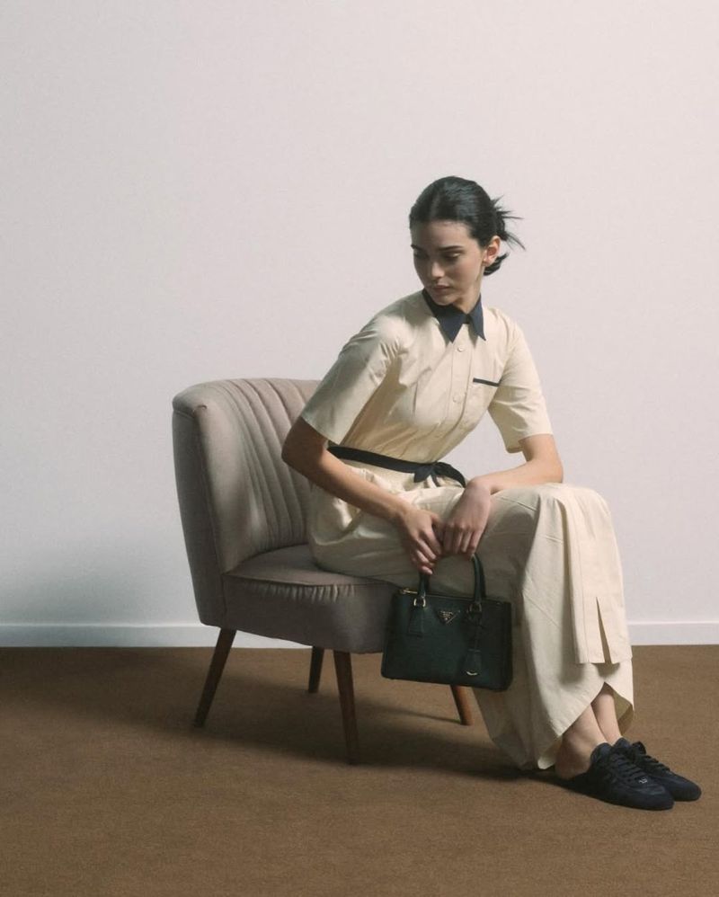 A woman sits in a beige chair holding a dark green purse, wearing a light beige dress, with a white wall and brown carpet behind her.