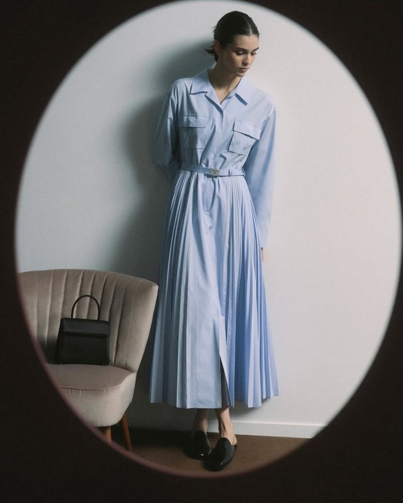 A slender woman in a light blue dress and black shoes standing in front of a white wall and light beige chair with a black bag.