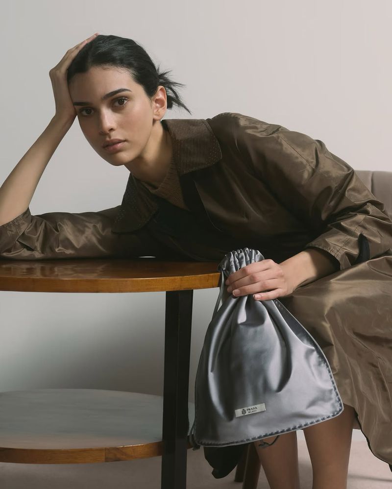 Portrait of a beautiful woman sitting at a table holding a gray drawstring bag in an indoor setting, wearing a brown jacket.