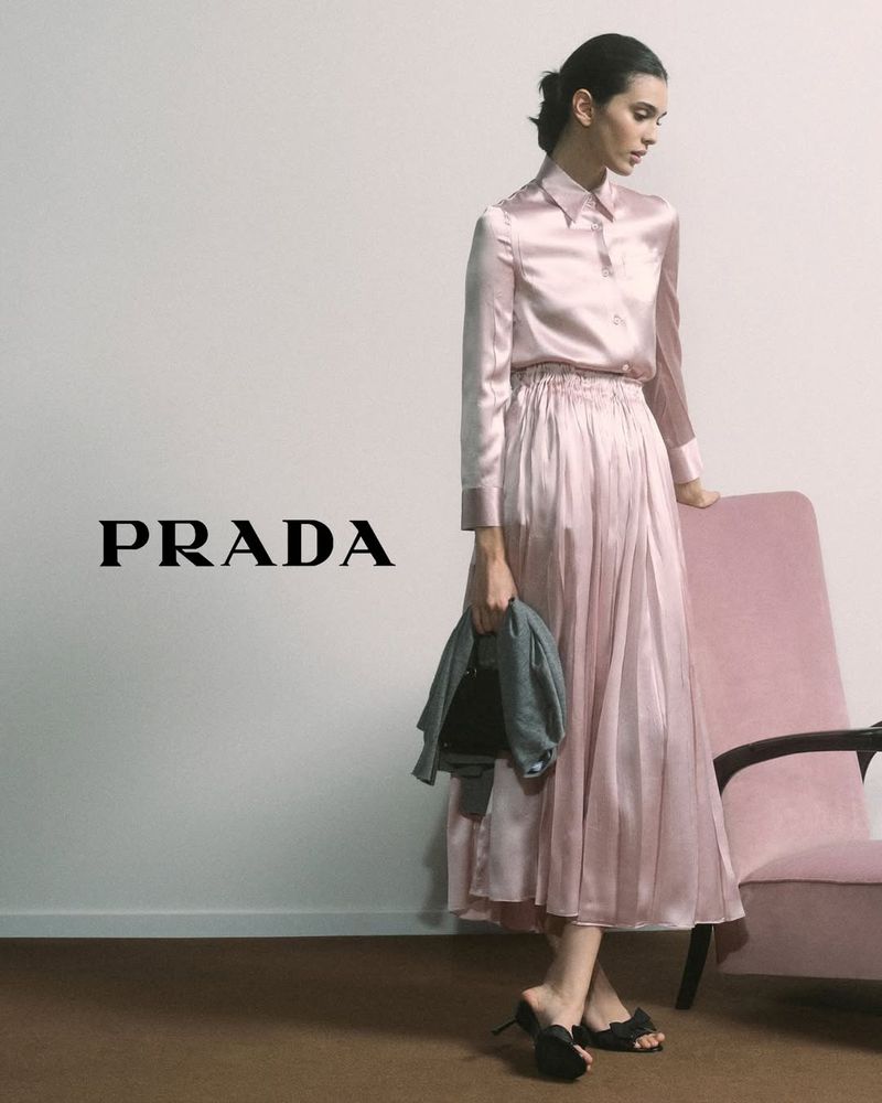 Woman in pink Prada satin shirt and skirt holding jacket, standing next to a pink chair in a fashion studio setting.