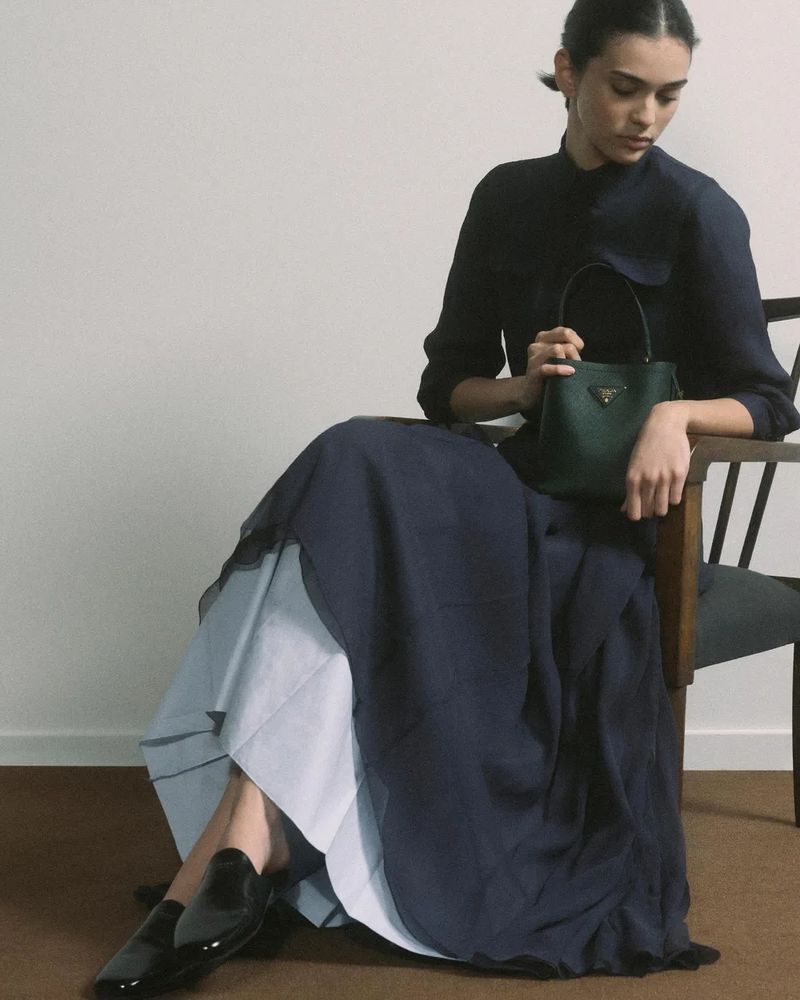 A woman in a long blue skirt and black blouse sits in a chair, holding a small green Prada handbag in a light-filled indoor setting.