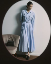 Model in a light blue belted pleated midi shirt dress standing against a white wall next to an armchair with a black handbag.