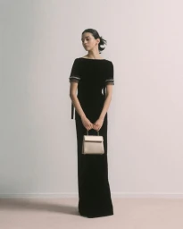 Model in a long black velvet dress holding a light-colored handbag against a plain background for Prada