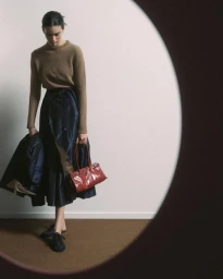 Model wearing a tan knit sweater and dark blue skirt holding a red handbag against a neutral studio backdrop