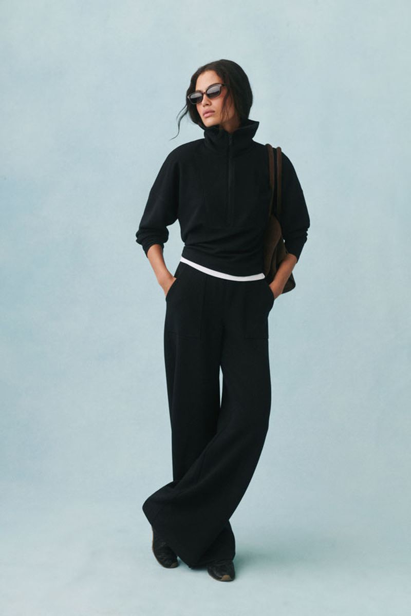 Woman in black turtleneck sweater and wide-leg pants poses against a light blue backdrop, accessorized with sunglasses and brown shoulder bag.