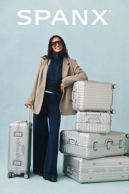 A smiling woman in a blazer and sunglasses stands by a stack of suitcases, promoting travel style for Spanx clothing brand.