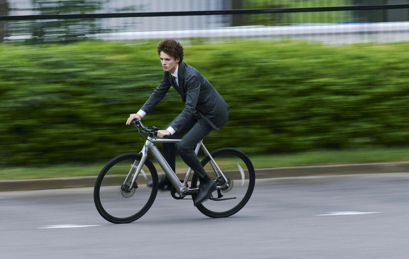 Young adult male in a suit rides a silver bicycle on a road with blurred green foliage in the background, captured in a motion-blurred image.