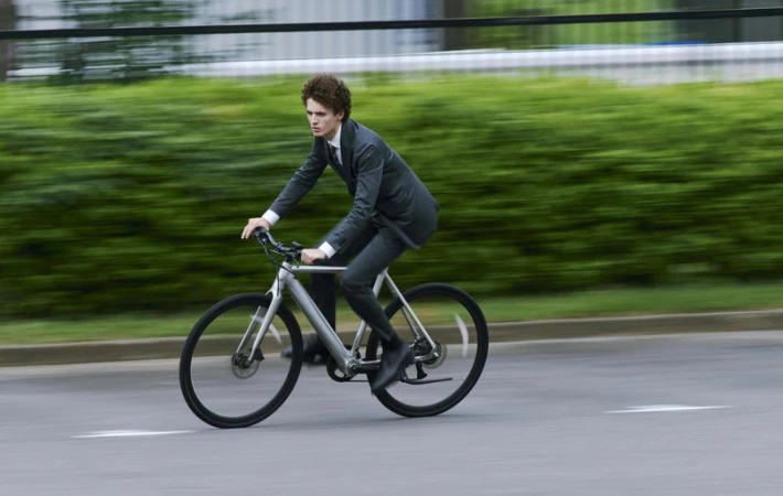 Young adult male in a suit rides a silver bicycle on a road with blurred green foliage in the background, captured in a motion-blurred image.