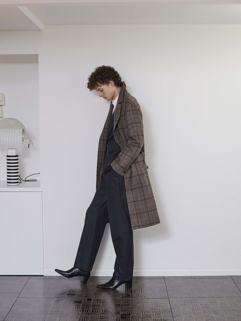 Young man in brown plaid overcoat, navy suit, and black heeled boots poses indoors against white wall near cabinet and striped lamp.