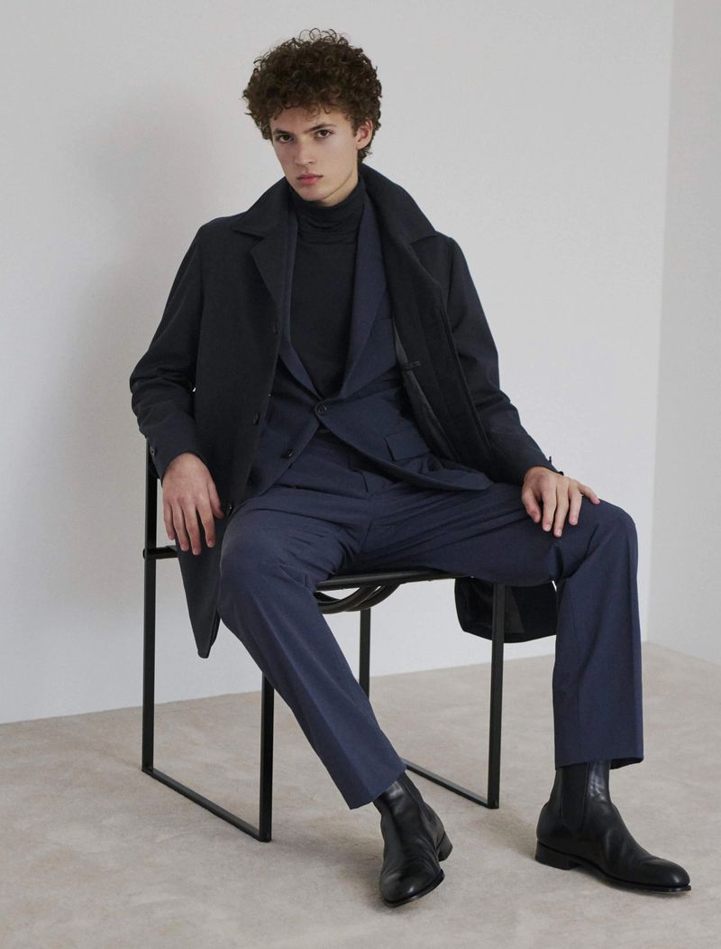 Man in navy suit and black coat seated, wearing black turtleneck and ankle boots, posing in a minimalist indoor studio.