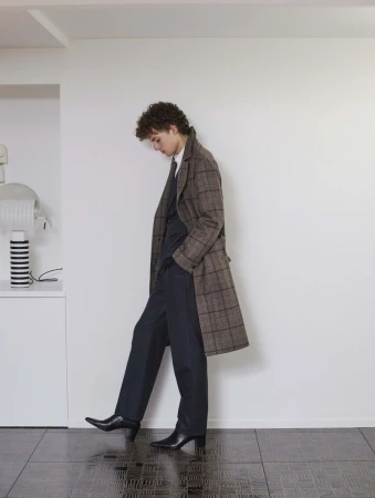 Model wearing a brown plaid coat over dark suit trousers and black boots against a plain white wall