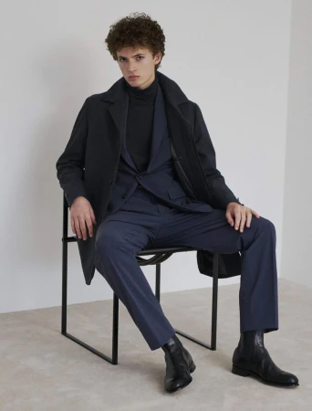 Model seated on a black chair wearing a dark coat, blue suit, and black boots against a plain wall.
