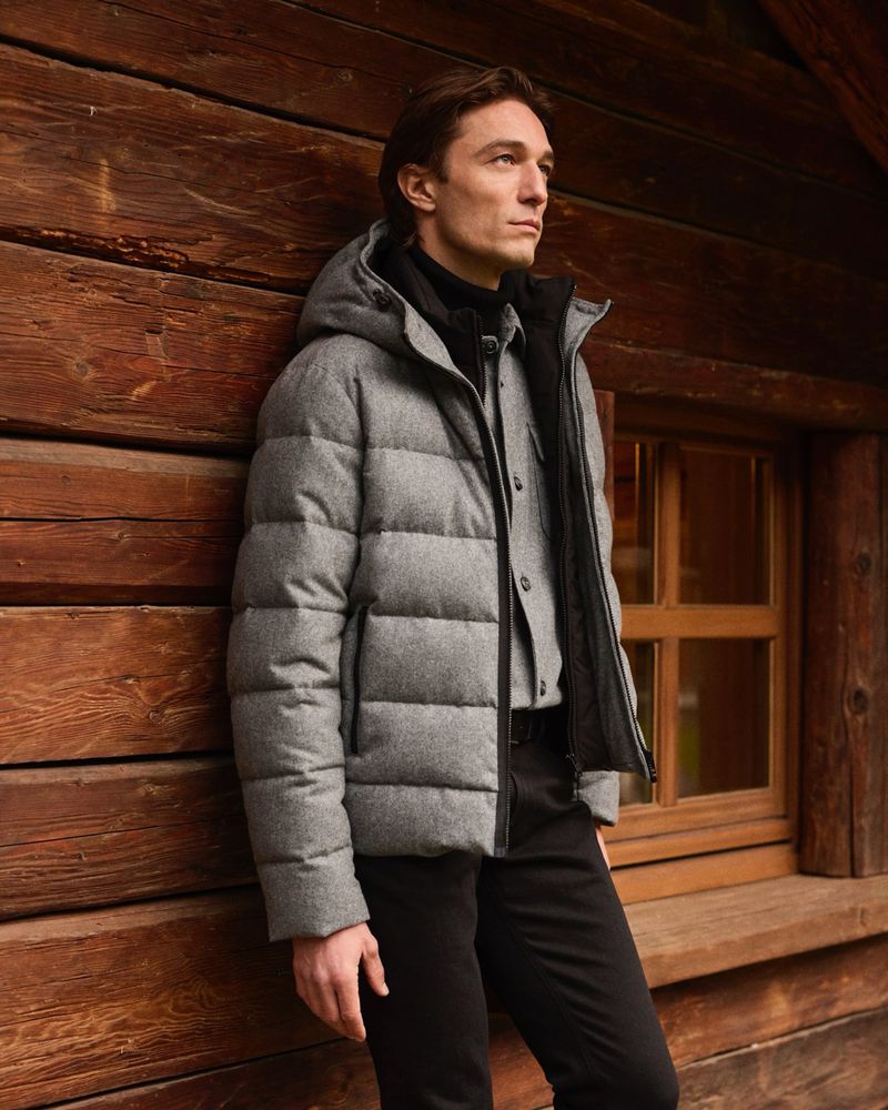 Man wearing a grey puffer jacket and black trousers leaning against a rustic wooden cabin wall.