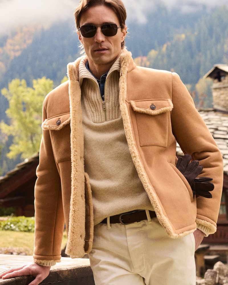 Man in shearling jacket, sunglasses posing outdoors with mountains in the background. Fashion portrait shot in natural light.