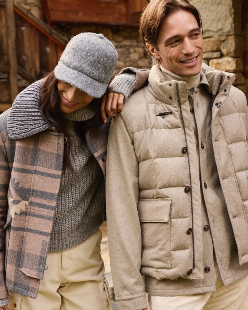 A smiling man and woman in winter clothing are photographed outside in a medium shot, wearing a puffer vest, jacket, and wool cap.