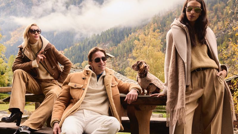 Three people pose outdoors with a dachshund dog wearing a coat, against a backdrop of mountains and autumn trees, all in earth-tone fashion.