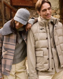 Two models wearing winter apparel including a plaid jacket and a quilted puffer vest against a stone background.