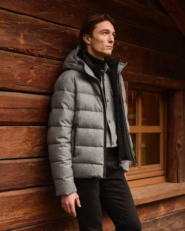 Man wearing a grey puffer jacket and black trousers leaning against a rustic wooden cabin wall.