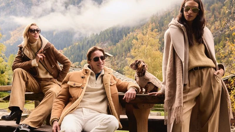 Three models wearing winter coats and sunglasses with a small dachshund dog on a wooden table outdoors against a mountain background.