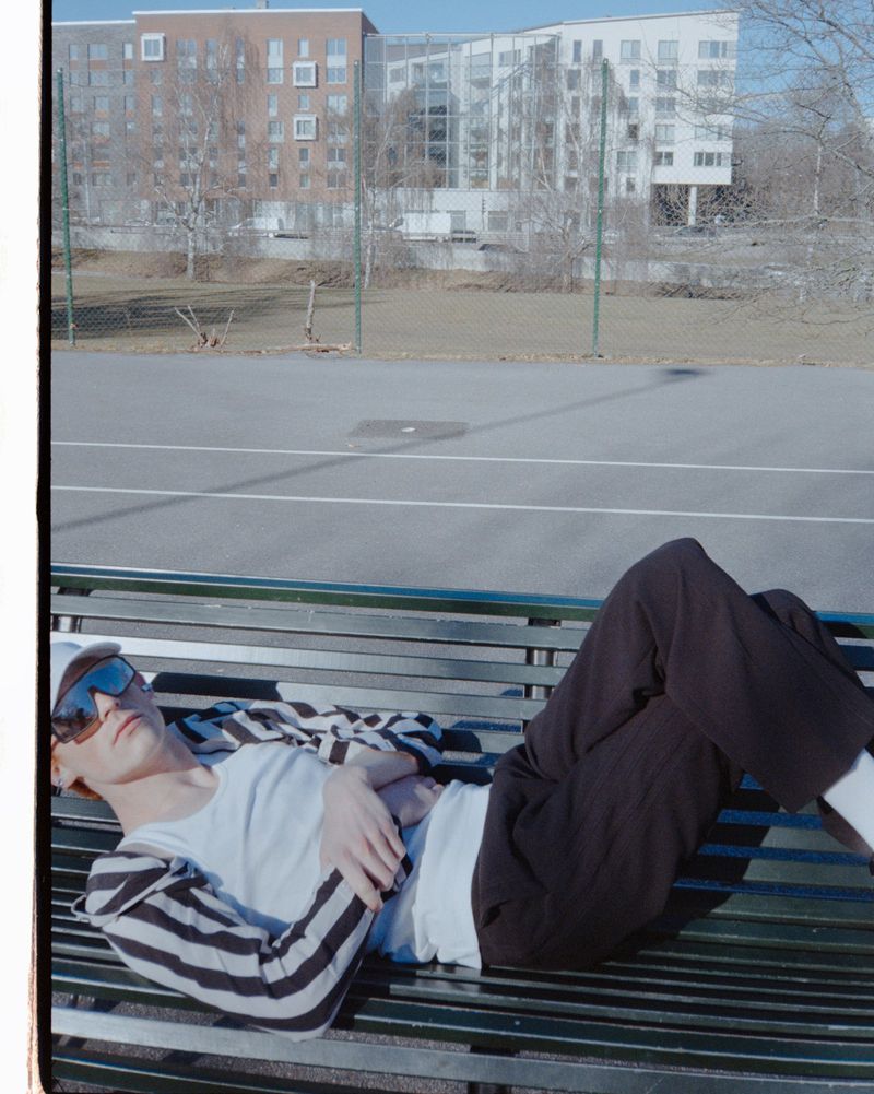 Young man in sunglasses lying on a park bench in sunlight, with a striped jacket and a modern apartment complex in the background.