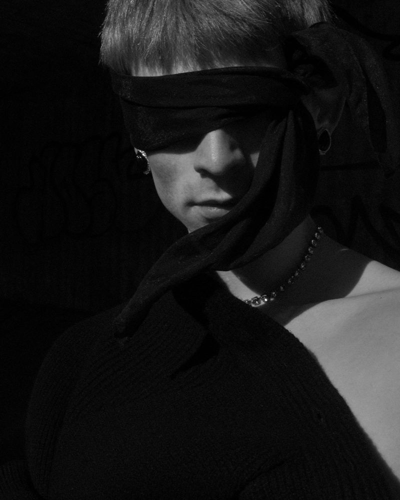 Portrait of a male model in black and white, wearing a black scarf as a blindfold, accessorized with a chain and earrings.