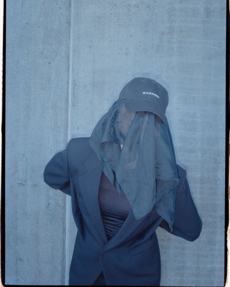 Person in blazer jacket, hat, and sheer fabric, posing against concrete wall in blue and gray tones. Fashion and style concept.
