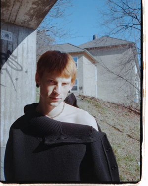 Young man with reddish-brown hair wearing a dark sweater outdoors near a concrete structure for Herdes Magazine editorial.