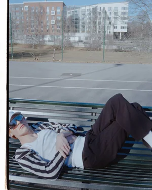 Man in dark sunglasses and striped jacket lying on a park bench outdoors