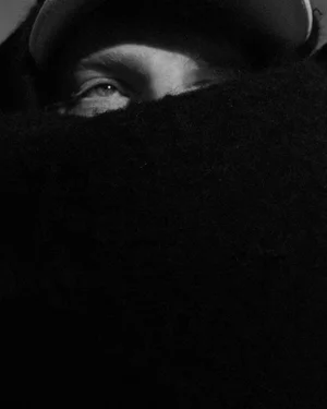 Close-up monochrome portrait of a person with a dark scarf wrapped over the lower face and a cap visible above.