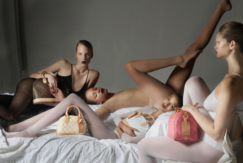 Fashion shot featuring four women posing on a bed with Louis Vuitton handbags in a studio setting, highlighting luxury fashion and style.