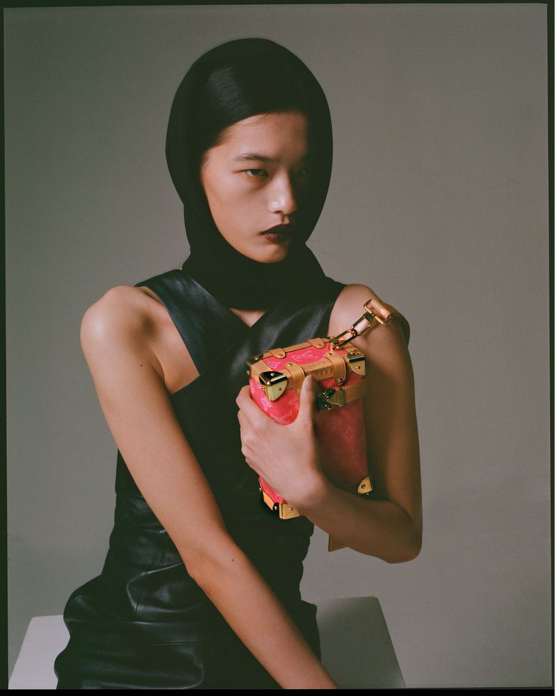 An Asian woman poses with a pink Louis Vuitton trunk bag in a studio setting, wearing a black head covering and a sleeveless dress.