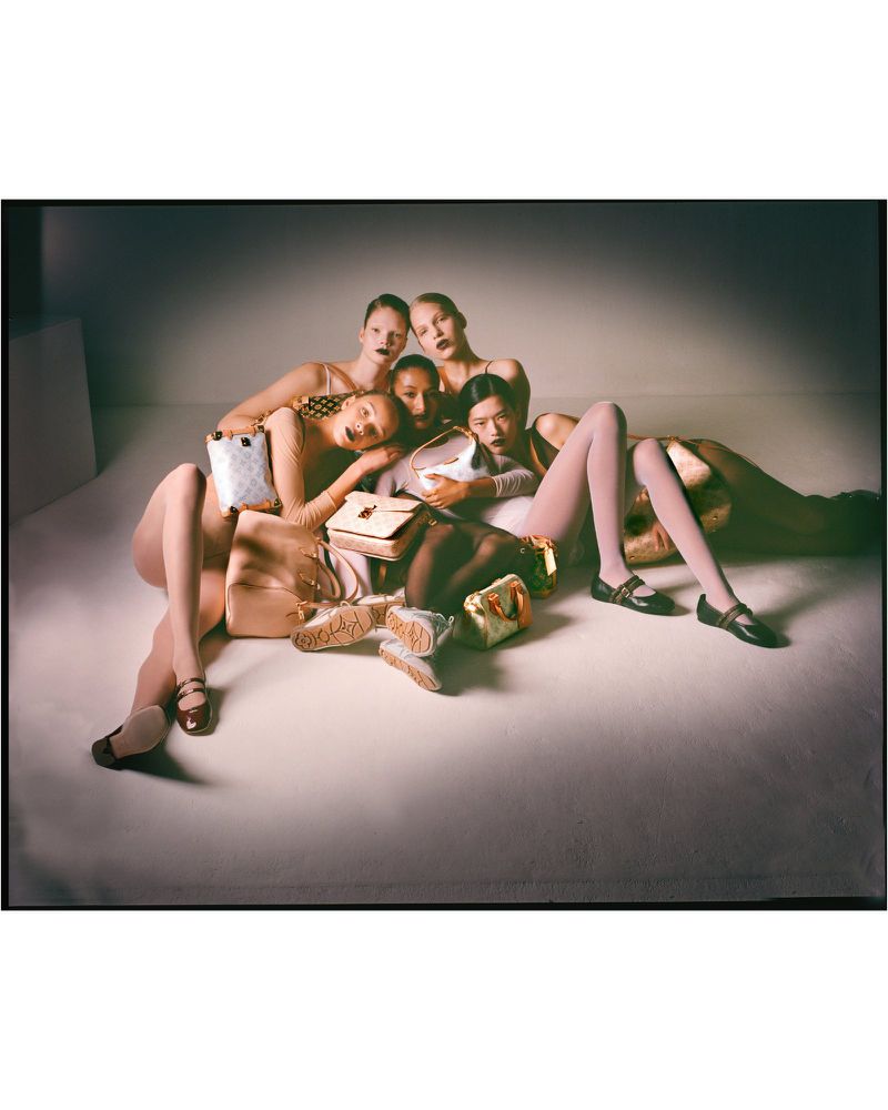 Group of fashion models poses with designer handbags in a photo studio. They wear various outfits and bold lipstick. Soft lighting.