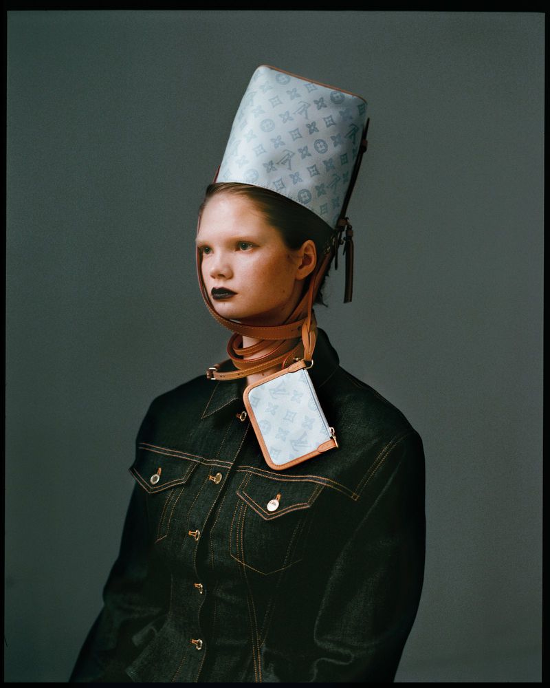 Fashion shot of a fair-skinned model wearing a Louis Vuitton hat, matching purse, and denim jacket in a grey studio setting.