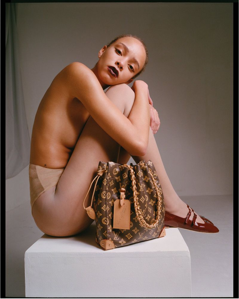 Adult female model in tights sits on a white cube next to a Louis Vuitton handbag and patent leather shoes in a studio setting.