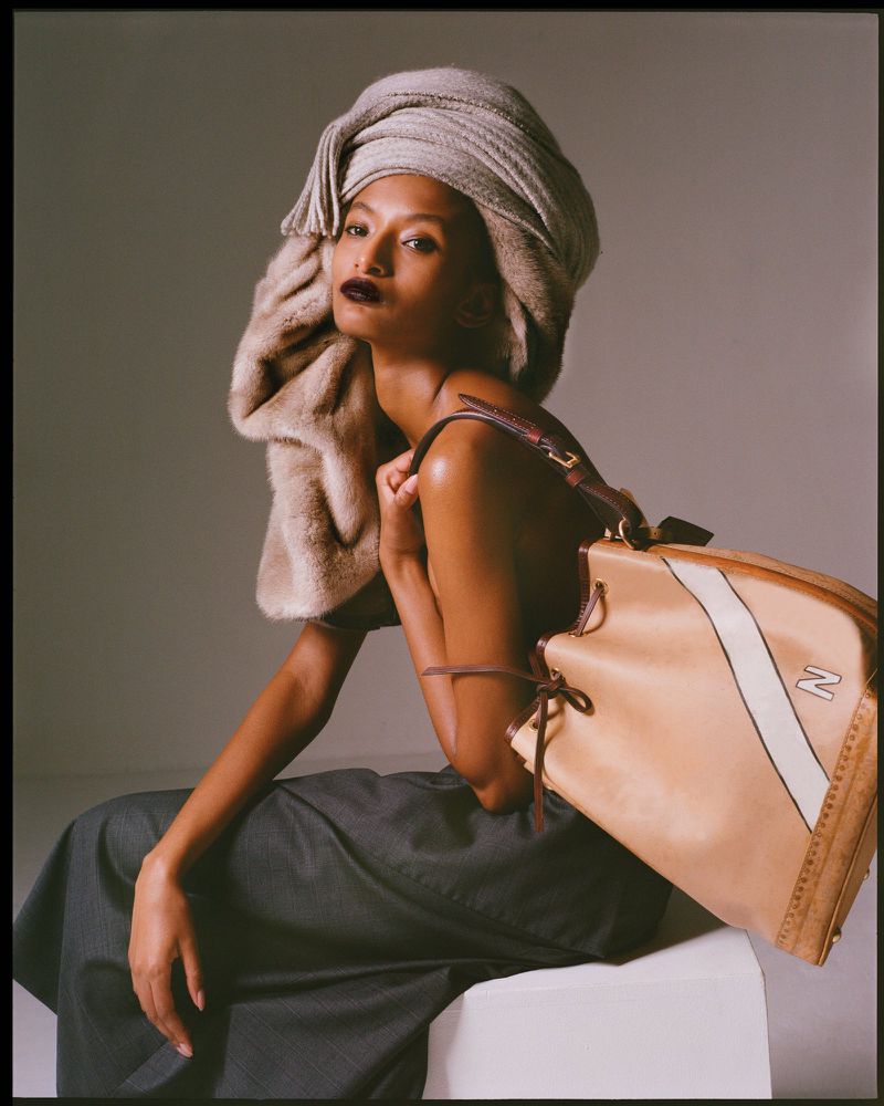 Fashion shot of a woman sitting and modeling a tan leather handbag with a scarf wrapped around her head in a studio with gray background.