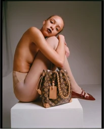 Model seated on a white block with monogram handbag and red shoes against a neutral studio backdrop